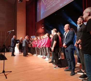 Na scenie grupa osób. Mężczyzna wręcza dyplomy. Na przodzie mikrofon