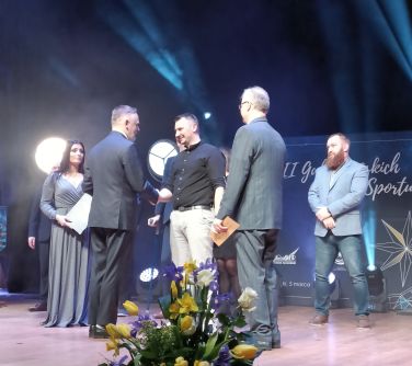 Scena. Mężczyzna gratuluje drugiemu. Obok po prawej stoi dwóch mężczyzn. Po lewej kobieta w długiej sukni