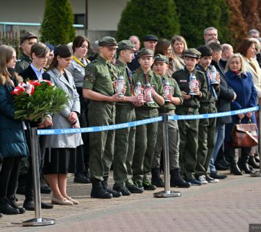 Dwór. Na chodniku za niebieską szarfą stoją osoby. Na przodzie harcerze trzymają znicze. Jedna kobieta trzyma wiązankę z...