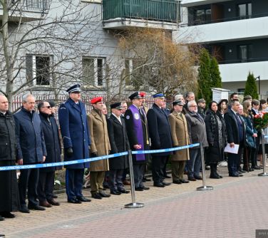 Dwór. Na chodniku za niebieską szarfą stoją osoby po cywilu, mundurowi, księża