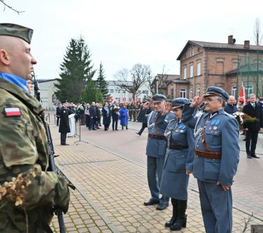 Dwór. Troje weteranów, kobieta i dwóch mężczyzn salutują przed pomnikiem. W tle osoby.