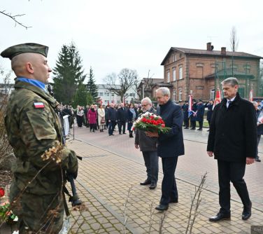 Dwór. Trzech mężczyzn składa wiązankę biało-czerwoną pod pomnikiem. W tle osoby.