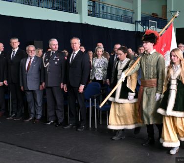 Aula szkolna. Osoby stoją przed krzesłami. Po środku maszeruje sztandar nastolatków przebranych w dawne stroje