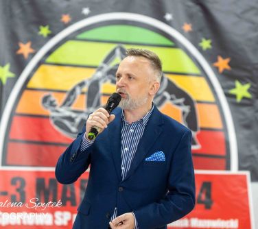 Mężczyzna w granatowej marynarce przemawia do mikrofonu. W tle kolorowy baner