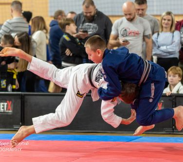 Hala sportowa. Dwóch chłopców w uchwycie w powietrzu walczą na macie. W tle widowania