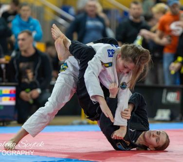 Hala sportowa. Dwie kobiety w uchwycie walczą na macie. Jedna, w ciemym kimonie leży na macie, druga w białym pochyla się...