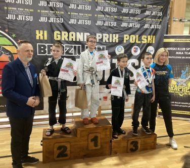 Hala sportowa. Dzieci pozują na podium z medalami i dyplomami w dłoniach. Po prawej kobieta w stroju sportowym. Po lewej...