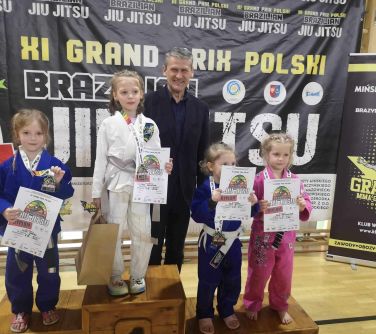 Hala sportowa. Czworo dzieci na podium, dwie dziewczynki na trzecim miejscu. Za nimi usmiechnięty mężczyzna w marynarce