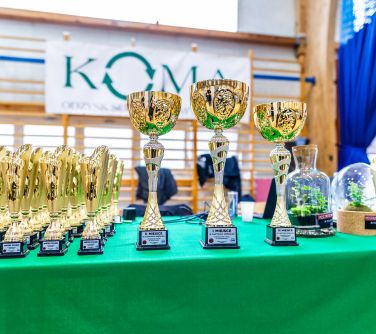 Hala sportowa. Stół przykryty zielonym obrusem. Zdjęcie pucharów różnej wielkości