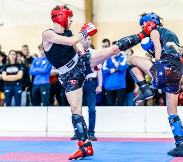 Hala sportowa. Na macie walczy dwóch mężczyzn w strojach sportowych i kaskach ochronnych. Jeden kopie drugiego w szyję.