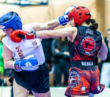 Hala sportowa. Dwie kobiety walczą w strojach sportowych, kaskach i rekawicach. Jedna drugą uderza rękawicą