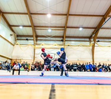 Hala sportowa. Na matach walczy dwóch mężczyzn. Mężczyźni w strojach sportowych, kaskach i rękawicach. Po lewej na krześle...
