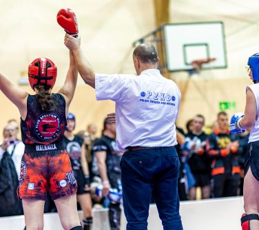 Hala sportowa. Na piankowej macie sędzia podnosi rękę kobiety po lewej stronie. W tle kibice