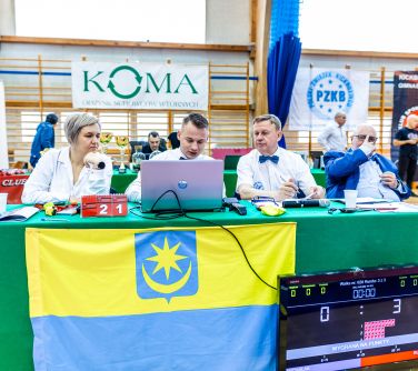 Hala sportowa. Przy stole przykrytym zielonym obrusem siedzą trzy osoby. Obok na krześle siedzi mężćzyzna i kucająca osoba...