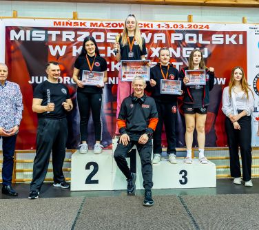 Hala sportowa. Na podium stoją osoby. Po bokach stoją po dwie osoby. Na przodzie na podium siedzi mężczyzna
