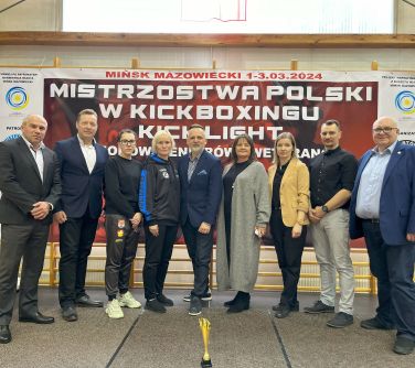 Hala sportowa. Dziewięć osób pozuje do zdjęcia. Przed nimi na podłodze stoi mały puchar