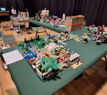 Pomieszczenie. Na stołach ustawionych w litere L, przykrytych zielonymi obrusami stoją budowle z lego