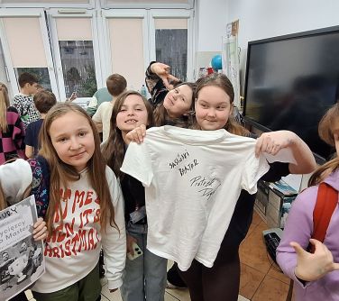 Sala lekcyjna. Pięć dziewczynek pozuje do zdjęcia. Jedna pokazuje jasną koszulkę z podpisami