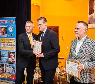 Aula. Trzech mężczyzn w marynarkach pozuje do zdjęcia. Dwóch z nich trzyma ramki ze zdjęciem