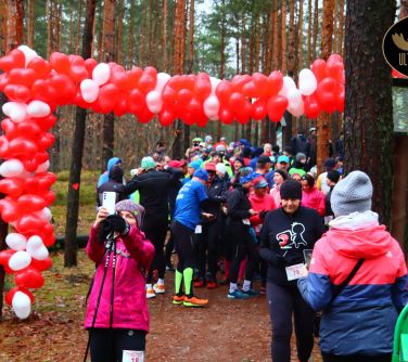 Ścieżka w lesie. Pomiędzy dzrzewami zawieszony łańcuch biało-czerwonych balonów wskazujący start biegu. Na starcie duża...