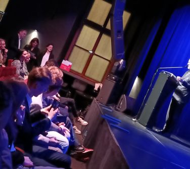 Pomieszczenie. Przed sceną na krzesłach siedzą nastolatkowie. Na scenie kawałek mównicy i zarys mężczyzny