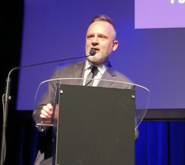 Pomieszczenie. Mężczyzna z bródką przemawia przy mównicy do mikrofonu