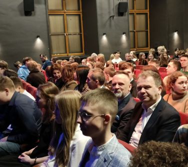 Pomieszczenie. Osoby siedzą w rzędach na fotelach
