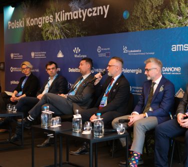 Siedem osób siedzi na krzesłach. Przed nimi dwa czarne stoliki ze szklankami i butelkami wody. Mężczyzna przemawia do...