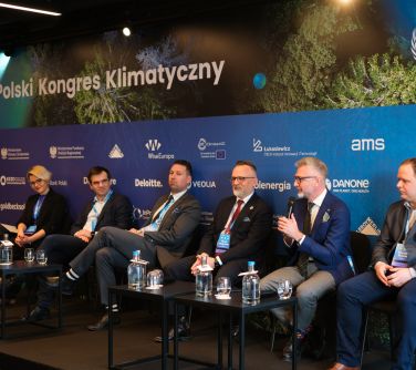 Siedem osób siedzi na krzesłach. Przed nimi dwa czarne stoliki ze szklankami i butelkami wody. Mężczyzna przemawia do...