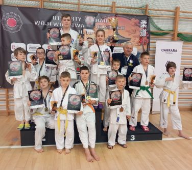 Hala sportowa. Trzynastka dzieci w białych kimonach stoi na podium prezentując dyplomy. Pomiędzy nimi mężczyzna.