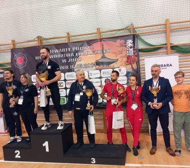 Hala sportowa. Dziewięc osób pozuje do zdjęcia. Cztery osoby na podium, reszta poza. Niektórzy z pucharami