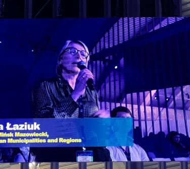 Widok na telebim, który przedstawia kobietę w okularach przemawiającą do mikrofonu
