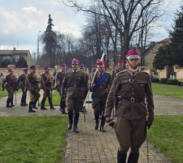 Grupa ułanów idzie chodnikiem z kostki. W rękach szable. Na głowie różowe czapki