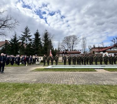 Na dworze, na placu. Szpaler żołnierzy i orkiestry. Na przodzie z lewej do mikrofonu przemawia mężczyzna