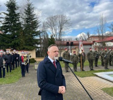 Na dworze. Mężczyzna w ciemnym płaszczu przemawia do mikrofonu. W tle orkiestra i żołnierze