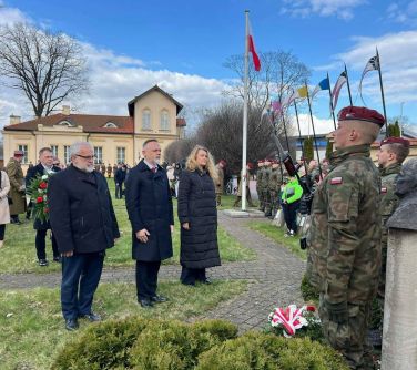 Na dworze dwóch mężczyzn i kobieta składają hołd przed pomnikiem. W tle osoby, żołnierze i budynek muzeum