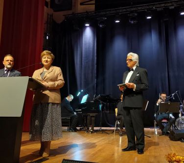 Pomieszczenie. Na scenie po lewej przy mównicy kobieta która przemawia, obok mężczyzna. Po prawej stoi mężczyzna w garniturze