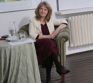 Pomieszczenie. W zielonym fotelu siedzi kobieta z założoną nogą na nogę. Obok stolik przykryty jasną płachtą, na którym...
