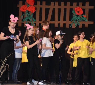 Pomieszczenie. Na scenie grupa osób występuje. Po prawej osoby w żółtych koszulkach