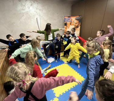 Pomieszcznie. Na kolorowych, piankowych matach kobieta z dziećmi stoją w rozkroku, trzymają dłonie wyciągnięte w bok