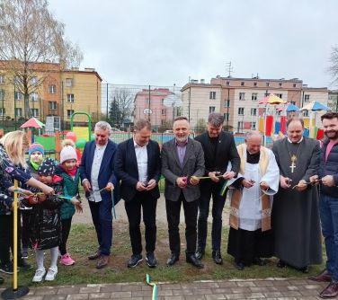 Na dworze. Kilku mężczyzn, ksiądz, kilkoro dzieci i kobieta przecinają wstęgę. W tle plac zabaw