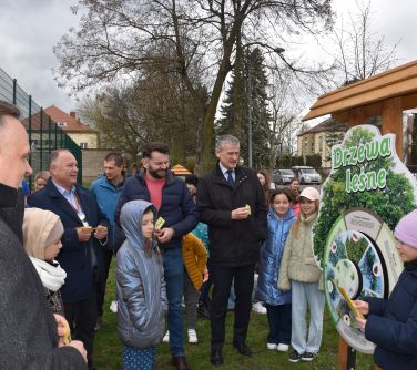 Na trawie stoi grupa osób dorosłych i dzieci. patrzą na stojak w kształcie drzewa z napisem "Drzewa leśne" z kołem fortuny.
