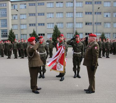 Na dworze. Na placu szpaler żołnierzy w czerwonych beretach. Na przodzie dwóch stojących na przeciw siebie salutujących,...
