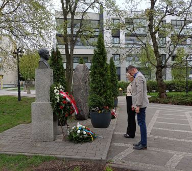 Kobieta i mężczyzna oddają hołd przed wysokim pomnikiem. Pod pomnikiem dwie wiązanki, jedna na stelarzu oparta o pomnik,...