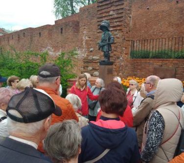 Na dworze. Grupa osób słucha opowieści kobiety. Grupa stoi przy postumencie młodego żołnierza. W tle barbakan