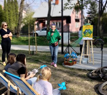 Na dworze, skwer. Kobieta w zielonej bluzie mówi do mikrofonu.  Na pierwszym planie dziewczynki siedzą na leżakach.