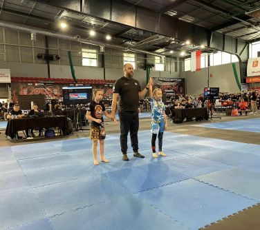 Hala sportowa. Na niebieskiej macie sędzia i dwie zawodniczki. Sędzia podnosi ręke jednej z nich. W tle trybuny.