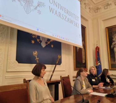 Pomieszczenie. Przy drewnianym stole siedzą cztery kobiety. Przed nimi mikrofony. Za kobietami na ścianie wisi ekran.