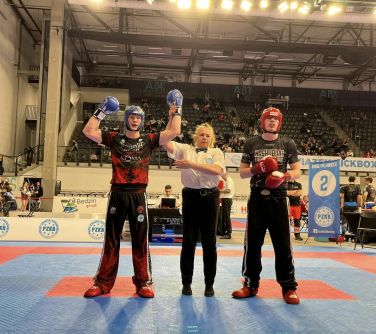Hala sportowa. Na macie sędzia i dwaj zawodnicy. Sędzia podnosi rękę jednego z nich. W tle trybuny.