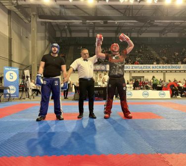 Hala sportowa. Na macie sędzia i dwaj zawodnicy. Sędzia podnosi rękę jednego z nich. W tle trybuny.